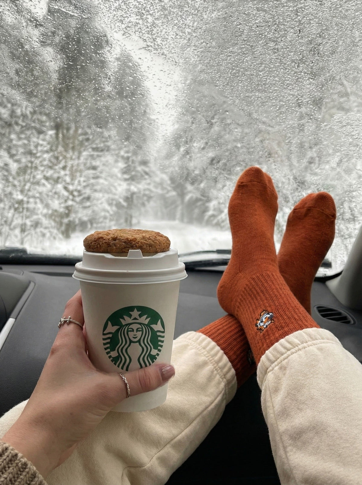 Füße in orangen Good Willy Socken im Auto vor verschneiter Landschaft, mit Kaffeebecher (KI-generiert)